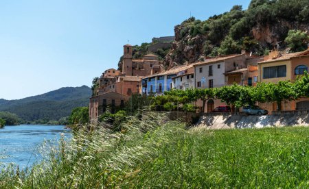 A serene river gently flows by a historic village with terracotta roofs, nestled against a backdrop of lush hills under a clear blue sky, capturing the essence of rural tranquility