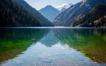 a serene alpine lake with crystal-clear waters reflecting the sky, bordered by dense coniferous forests and a backdrop of snow-dusted mountains under a blue sky