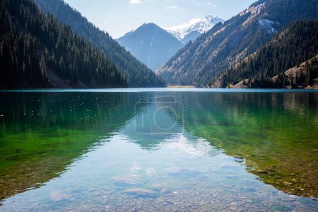 a serene alpine lake with crystal-clear waters reflecting the sky, bordered by dense coniferous forests and a backdrop of snow-dusted mountains under a blue sky