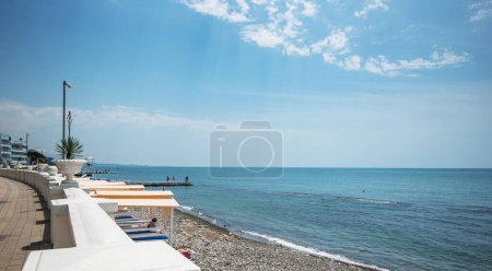 A serene pebble beach with sunbeds lined along a promenade, a clear blue sky above, and a calm sea inviting relaxation.