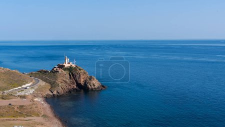 lighthouse perched on a rocky promontory extending into the sea, with a road winding towards it against the backdrop of a vast, calm blue ocean under a clear sky.