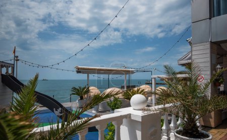 A coastal terrace with string lights, sun umbrellas, and loungers, offering a view of a calm sea and a distant ship under a partly cloudy sky, suggests a relaxed, seaside ambiance.
