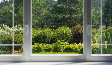 Through the panes of a window, reveals a meticulously cared for garden, resplendent with a variety of shrubs and blooming flowers, and flanked by tall trees, offering a slice of nature's tranquility.