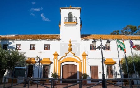 a traditional Spanish building with a white facade and distinctive yellow architectural details, featuring a bell tower, under a clear blue sky, flanked by the Spanish and Andalusian flags