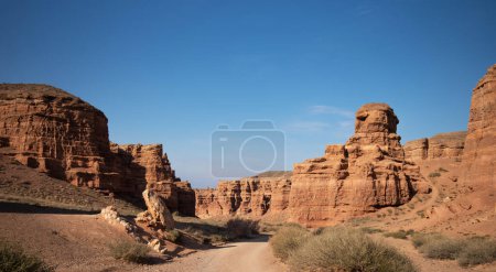 A rugged dirt road leads through an awe-inspiring canyon with towering red sandstone cliffs under a clear blue sky, highlighting the natural architecture and grandeur of the desert