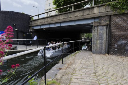 Photo for London - 05 29 2022: Boat with people sitting cruising under Harrow RD bridge on Paddington Basin, with a dog looking on. - Royalty Free Image