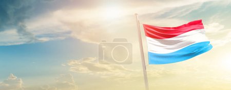 Téléchargez les photos : Luxembourg waving flag in beautiful sky with sun - en image libre de droit