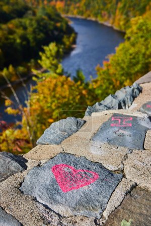 Foto de Imagen de Detalle de pared de piedra con tiza de corazón rosa y río suave con bosque de otoño detrás - Imagen libre de derechos