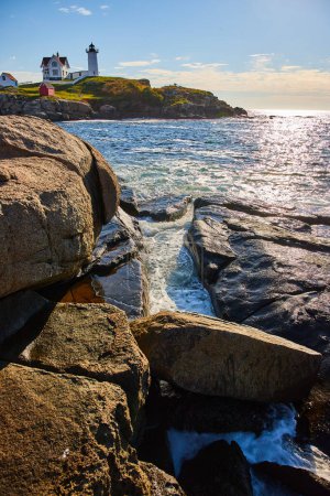 Téléchargez les photos : Image des côtes rocheuses du Maine avec des crevasses remplies de vagues océaniques et un phare en arrière-plan - en image libre de droit
