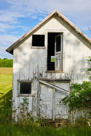 Photo for Image of Vertical shot of abandoned farmhouse in grassy field near farmland with busted windows and door - Royalty Free Image
