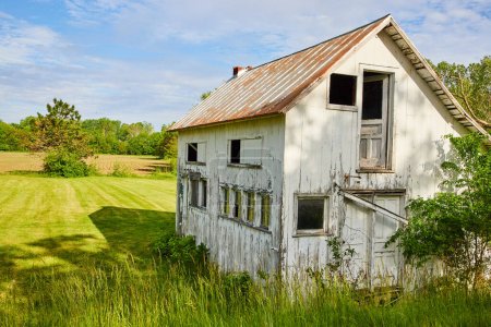 Image d'une ferme abandonnée dans un champ herbeux près d'une terre agricole avec fenêtres et porte cassées