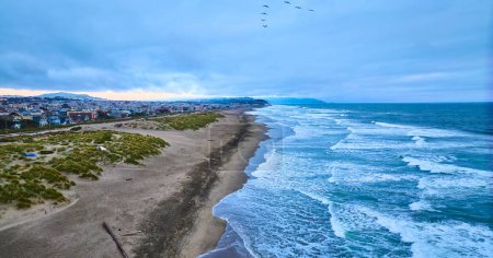 Téléchargez les photos : Image de basse altitude de vagues océaniques sans fin avec rivage sablonneux et oiseaux au-dessus et une ville sur la gauche - en image libre de droit