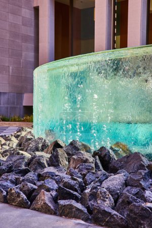 Photo for Image of Circular wall with distorted glass from falling water over large black rocks in front of building - Royalty Free Image
