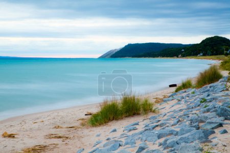Téléchargez les photos : Une plage tranquille à l'heure bleue dans un endroit côtier isolé. Des eaux douces, une plage de sable fin et une barrière rocheuse texturée créent une atmosphère sereine. - en image libre de droit