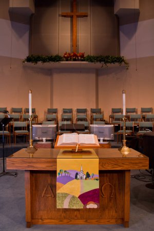 Photo for Tranquil Worship: A serene church interior in Fort Wayne, Indiana, showcases a wooden pulpit adorned with a colorful landscape, an open Bible, and elegant candles. A large wooden cross, accented by a - Royalty Free Image