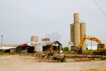 Photo for Symbolic of change and decay, a yellow CAT excavator demolishes a derelict building in Evansville, Indiana. Amidst the ruins, towering silos stand as a reminder of the areas industrial past. - Royalty Free Image