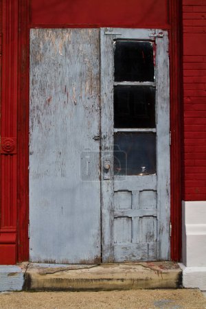 Téléchargez les photos : Charme urbain négligé : Une porte bleue grise altérée avec peinture pelante et vitre cassée se détache contre un mur de briques rouge audacieux à Allegan, Michigan. Le matériel usé ajoute à la nostalgie - en image libre de droit