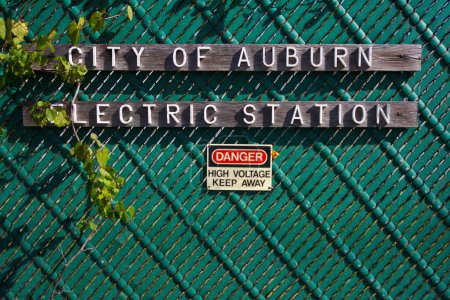 Photo for Close-up of weathered City of Auburn Electric Station sign on a vibrant teal chain-link fence, entwined with lush green vines, warning of high voltage dangers - Royalty Free Image