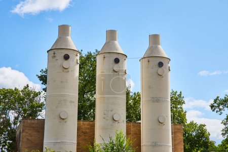 Téléchargez les photos : Imposante tour de silos industriels de couleur crème contre un ciel bleu vibrant, entourée d'un complexe industriel dans les jardins botaniques, Elkhart, Indiana, dépeignant l'harmonie entre l'industrie et la nature. - en image libre de droit