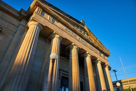 Téléchargez les photos : Golden hour sunrise illumine la grande architecture de la bibliothèque publique Muncie, Indiana, mettant en valeur ses colonnes ornées et sa maçonnerie sur un ciel bleu clair. - en image libre de droit
