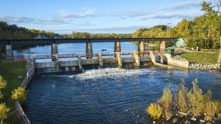 Foto de Hora dorada sobre el río Hurón, Michigan mostrando una serena cascada desde la presa de Argo, un puente ferroviario y actividades recreativas. - Imagen libre de derechos