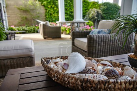Photo for Coastal themed tranquil patio setting with a basket of seashells on a wooden table, comfortable wicker furniture and lush greenery in a Syracuse, Indiana home designed s, 2015. - Royalty Free Image