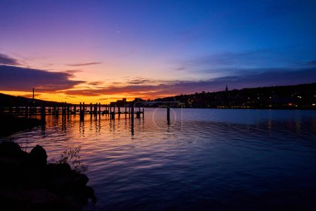 Serene autumn sunrise at Houghton, Michigan with a wooden dock extending into placid water, under a vibrant blue hour sky with a crescent moon