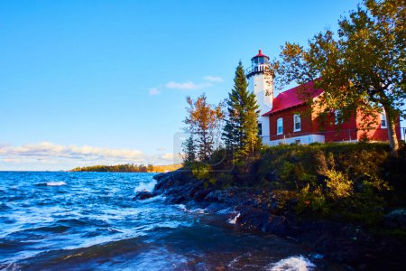 Malerischer Blick auf den historischen Eagle Harbor Leuchtturm am Lake Superior, Michigan, vor einem klaren blauen Himmel und schroffen Klippen, die maritime Sicherheit und Reisen repräsentieren.