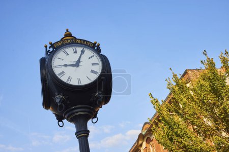 Historic Ypsilanti ornate street clock against a clear blue sky with an old brick building in the background, embodying tradition and history in Michigan.
