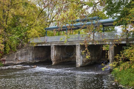 Concrete bridge with blue-roofed shelter over tranquil river amid autumn foliage in Homer, Michigan