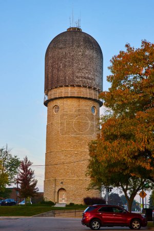 Golden hour sunlight illuminating the historic Ypsilanti Water Tower in Michigan, with an autumnal backdrop and a contrasting modern red car parked nearby.