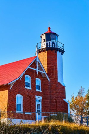 1871 Eagle Harbor Lighthouse in Michigan, a red-brick beacon perched on a grassy coastal bluff under a clear blue sky, framed by fall foliage.