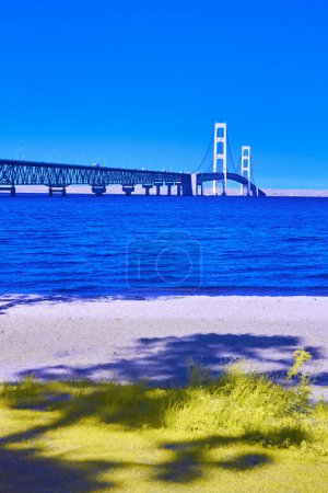 Fall 2017 view of the majestic Mackinac Bridge in Michigan, with its white pillars and cables, over vibrant blue water from a sandy beach.
