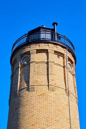 Photo for Classic yellow-brick lighthouse with a black metal rooftop, standing tall against a vibrant blue sky in Mackinac, Michigan. Symbol of maritime navigation and coastal tradition. - Royalty Free Image