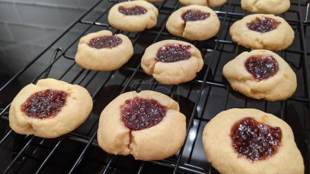 Téléchargez les photos : Biscuits fraîchement cuits à l'empreinte de pouce refroidissant sur un support métallique dans une cuisine confortable, remplie de confiture rouge scintillante - en image libre de droit
