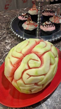 Brain-etched watermelon on vibrant plate, set in a Halloween themed kitchen with creatively decorated cupcakes in the background