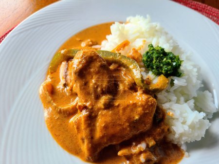 Herzhaftes Curry-Gericht mit Huhn und Paprika, serviert mit weißem Reis und grüner Kräutersoße, bei Tageslicht auf einem Holztisch, Fort Wayne, Indiana, 2023