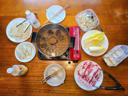 Overhead view of an Asian hot pot meal setup with simmering broth, raw ingredients and utensils on a wooden table, depicting communal dining and home cooking in Fort Wayne, Indiana, 2022.