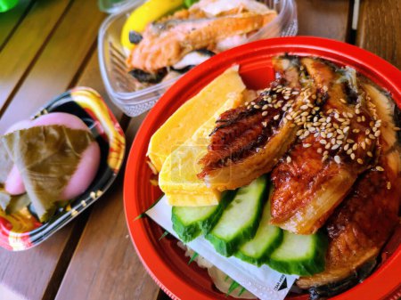 Appetitliche japanische Bento-Box mit gegrilltem Aal, Tamagoyaki und Gurkenscheiben, serviert mit Lachs und traditionellen Mochi-Süßigkeiten in einem lässigen Speisesaal in Columbus, Ohio, 2023