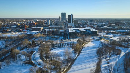 Winters Dawn Over Fort Wayne - Aerial view of a snow-kissed urban park, the frozen St. Marys River, and vibrant downtown cityscape in Indiana.