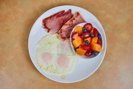 Traditionelles amerikanisches Frühstück mit sonnenbeschienenen Eiern, gebratenem Schinken und frischem Obst auf einer gemütlichen Tischplatte in Fort Wayne, Indiana.