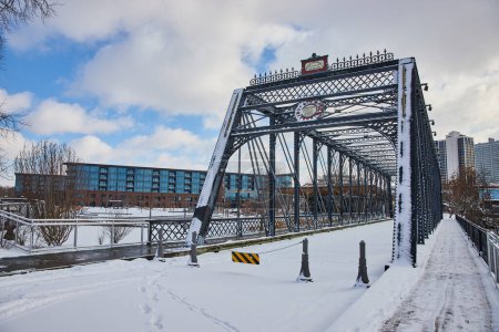 Winter scene at Promenade Park, Fort Wayne, Indiana featuring snowy Wells Street Bridge blending historical charm with modern cityscape.