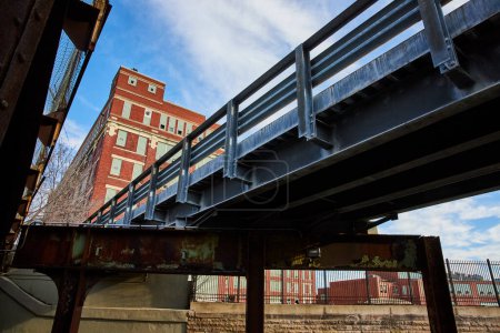 Industrial meets historic in Fort Wayne, Indiana - a sturdy, weathered bridge forefronts classic red brick architecture under a clear sky.