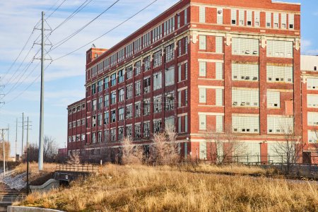 Edificio industrial abandonado de principios del siglo XX con ventanas rotas en medio de hierba seca, bajo cielo azul claro - Electric Works, Fort Wayne, Indiana