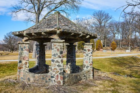Vintage stone gazebo in Lindenwood Cemetery, Fort Wayne, Indiana, bathed in sunlight amidst a serene park setting.