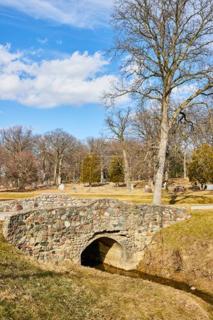 Stone bridge spanning a tranquil creek in Lindenwood Cemetery, Fort Wayne, Indiana. A picturesque scene of serenity and history in late fall.