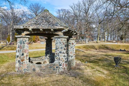 Antiguo pozo de piedra en medio de un paisaje sereno en el cementerio de Lindenwood, Fort Wayne, Indiana, que muestra la transición estacional y el encanto rústico.