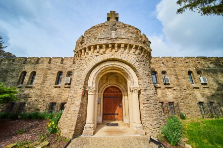 Obispo Simon Brute College torre de piedra de estilo medieval bajo el cielo parcialmente nublado de Indiana, con una cruz y puerta de madera ornamentada.
