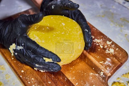Manos expertas en guantes negros amasando masa amarilla en una tabla de cortar bien utilizada, capturando el arte de hacer pasta en Fort Wayne, Indiana.