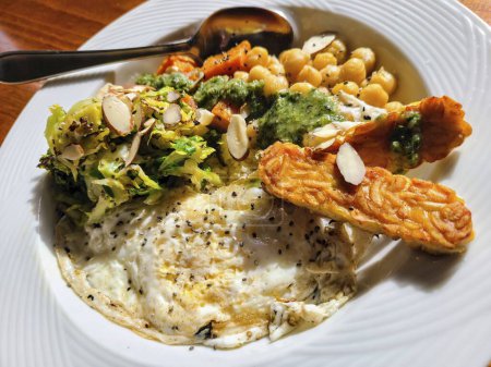 Gourmet vegetarian plate with crispy tempeh, runny egg, shaved brussel sprouts, almond slices, carrots, and chickpeas, served in a cozy indoor setting.